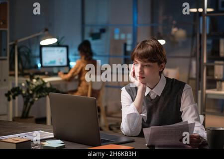 Young woman sitting at her workplace looking at laptop screen and working with documents during her deadline at office Stock Photo