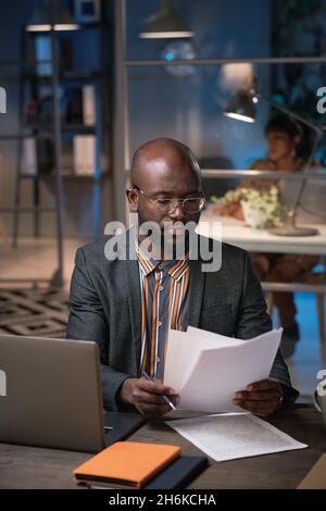 African businessman concentrating on his paper work, he sitting at the table in front of laptop and reading documents during his work at office till late Stock Photo