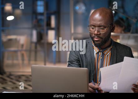 Serious African businessman working on laptop and examining documents while working late at office Stock Photo