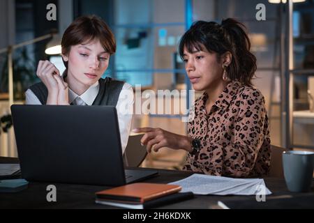 Businesswoman pointing at the screen of laptop and discussing online presentation with her colleague, they working at office till late evening Stock Photo