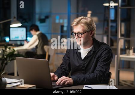 Young businessman in eyeglasses sitting at the table and working online, he typing on laptop while working late at office Stock Photo
