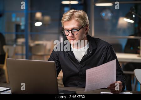 Young businessman in eyeglasses using laptop for typing contract while working at his workplace till late night at office Stock Photo