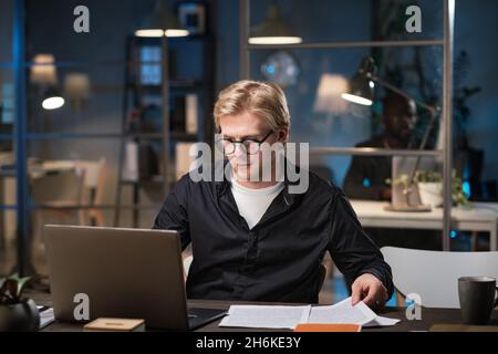 Young businessman in eyeglasses using laptop for online work, he working with document till late evening at office Stock Photo