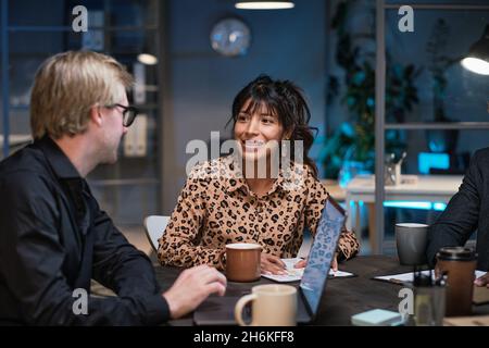 Young businesswoman sitting at the table working with documents and discussing with colleagues during a meeting till late evening at office Stock Photo