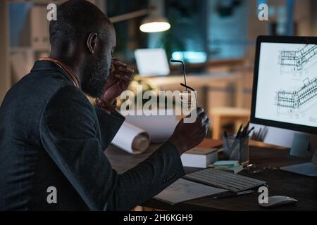 Tired African architect sitting at the table in front of the computer monitor with blueprint while working at office till night Stock Photo