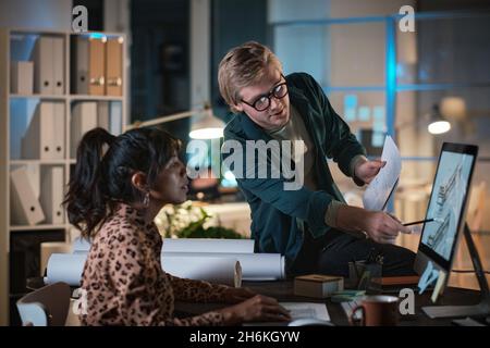 Young designer pointing at computer monitor and discussing blueprint with his colleague during their teamwork at office till late night Stock Photo