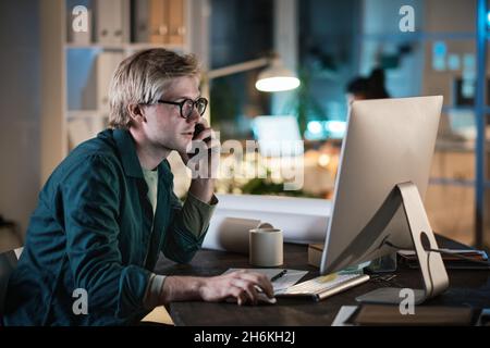 Young businessman in eyeglasses working on computer and talking on mobile phone while sitting at the table and working late at office Stock Photo