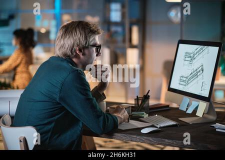 Young designer in eyeglasses sitting at the table and looking at computer monitor, he developing new project at office till late evening Stock Photo