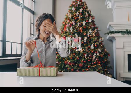 Excited young woman unpacking Christmas gift while sitting in room decorated with bright Xmas tree Stock Photo