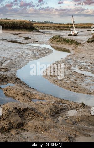 Small yacht aground on mud flats with narrow steam of water in the foreground Brancaster Staithe Harbour North Norfolk England Stock Photo