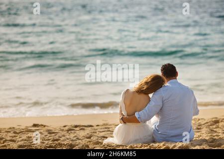 Newlyweds sitting close together at sunset on the beach in the sand, gazing out to sea. Bride and groom embrace each other near ocean and looking on s Stock Photo