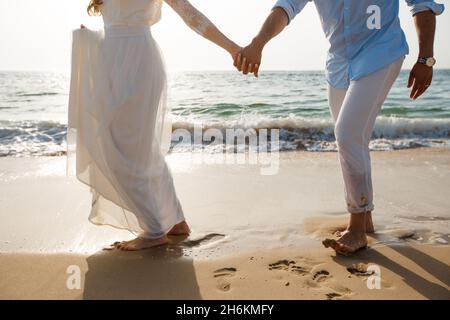 Newlyweds honeymoon. Barefoot bride in white dress and groom in white pants walking on romantic ocean beach. Young couple foots on golden sand. Stock Photo