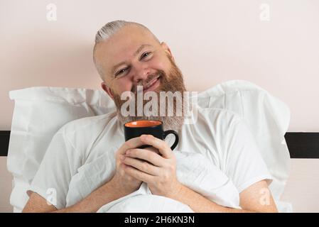 Happy bearded man enjoying morning coffee in bed. Smiling hipster guy starting the day. Closeup. Stock Photo