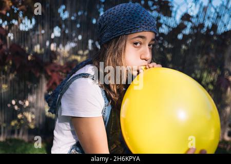 Teenage girl with a headscarf and long brown hair blowing up a huge yellow balloon sitting in a green garden on a sunny day. Adolescence Stock Photo