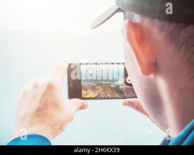 A guy in a cap takes photo from a phone with cracked screen. Shockproof phone. Focus on the phone. The head is out of focus. Stock Photo