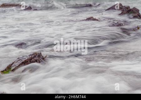 Soft blurred seawater flows around dark coastal rocks at Dollar Cove, Gunwalloe, Cornwall. Abstract, atmospheric scene in muted natural light Stock Photo
