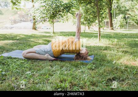 Portrait of an elderly woman sitting on a yoga mat and doing sports in the park. Stock Photo