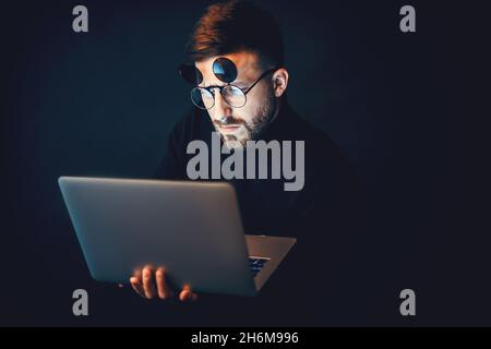 Young bearded hipster man wearing glasses while using laptop over black studio background  Stock Photo
