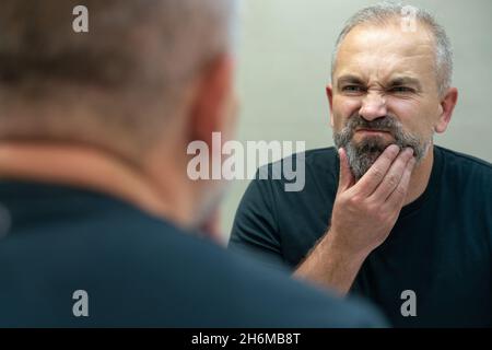 Middle-aged handsome man looking in mirror in bathroom touching his beard thinking of cutting it off Stock Photo