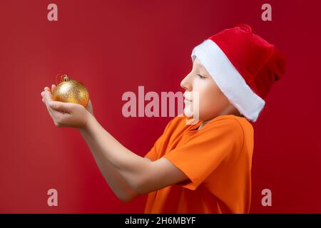 a boy in a New Year's Santa Claus hat stands sideways and holds a ball on the Christmas tree in his palms Stock Photo