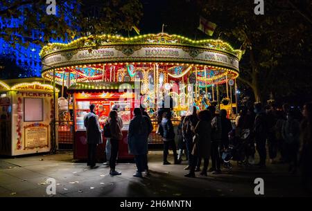 People stand by a brightly lit traditional funfair carousel and candy floss stall at night on the South Bank, London, UK, 14 November 2021