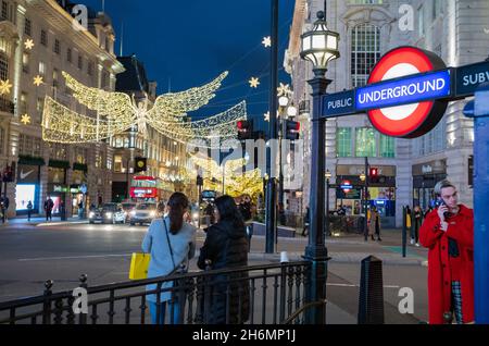 A yound man in a red coat speaks on the telephone at the entrance to Piccadilly Circus Tube Station as Christmas Lights illuminate the night streets Stock Photo