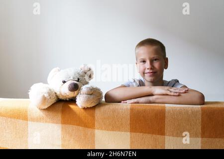 The boy plays with a teddy bear and looks out from behind the sofa. Happy caucasian child looking at the camera on the background of a light wall at home. Child leisure concept at home Stock Photo