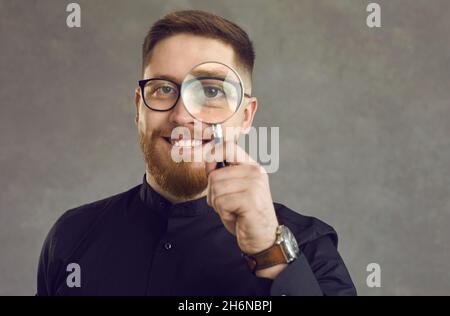 Positive young detective man looking through a magnifying glass while standing on gray background. Stock Photo
