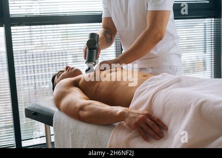 Medium shot of unrecognizable professional male masseur receiving chest muscles using massage gun percussion tool of muscular athlete man Stock Photo