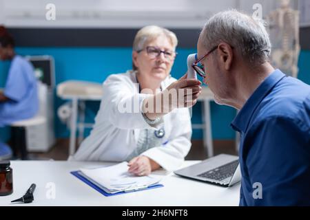 Physician using thermometer to measure temperature of aged patient, checking for flu symptoms. Woman doctor measuring fever of ill man using medical instrument at annual checkup visit. Stock Photo