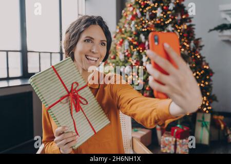 Young Italian woman online blogger making selfie with christmas gift on background of Xmas tree Stock Photo