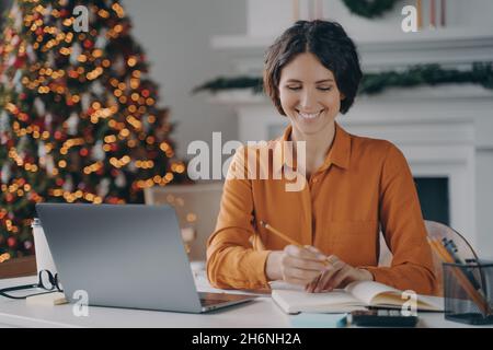Smiling italian female making notes, using laptop at home office during christmas holidays Stock Photo