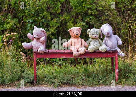 Four different coloured stuffed animals, teddy bears sitting on a red bench, one alone, Gotland, Sweden Stock Photo