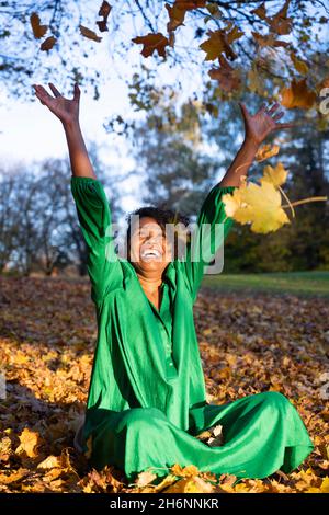 Portrait of a laughing, dark-skinned woman with curls in a green dress, sitting and throwing autumn leaves, outdoor shot in autumn, Germany Stock Photo