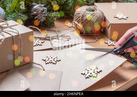 Packaging of ecological Christmas gifts - hand cutting recycled paper with scissors on the table with artificial fir tree with cones, envelope, twine, Stock Photo