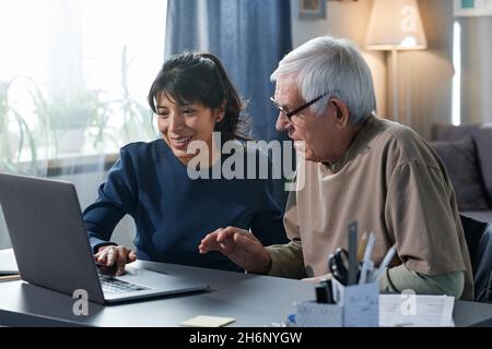 Friendly volunteer teaching to use the computer to senior man while they sitting at the table at home Stock Photo