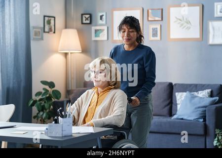 Woman caring about senior woman at home, she carrying her on a wheelchair to the table with laptop Stock Photo