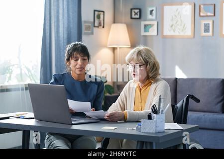Senior woman sitting at the table at home and learning to pay for bills online using her laptop with the help of volunteer Stock Photo