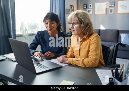 Young volunteer showing to senior woman how to use computer while they sitting at the table in the living room Stock Photo