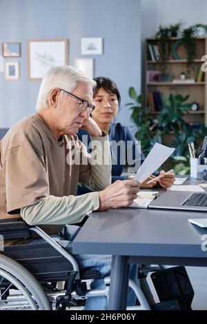 Senior man with disability reading a document at the table with laptop, he studying together with his teacher at home Stock Photo