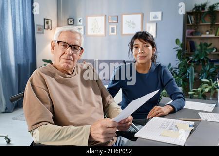 Portrait of senior man in eyeglasses looking at camera while reading documents at the table together with his assistant Stock Photo