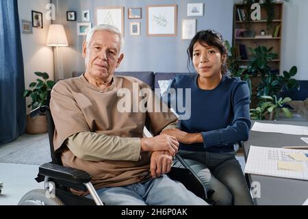 Portrait of senior man using wheelchair looking at camera while sitting in the room together with nurse Stock Photo