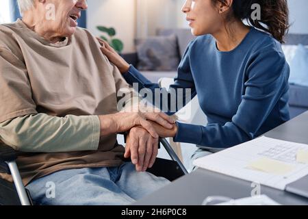 Close-up of senior man using wheelchair talking to the nurse while she holding his hands at the table Stock Photo