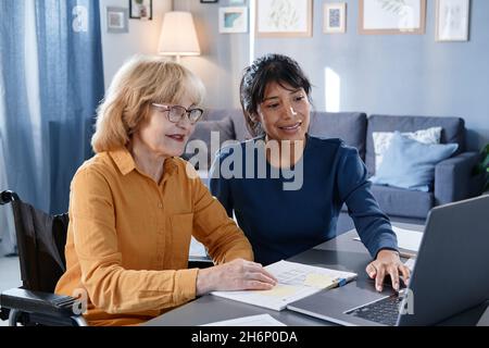 Senior woman in eyeglasses sitting at the table with documents and using laptop together with assistant in the room Stock Photo