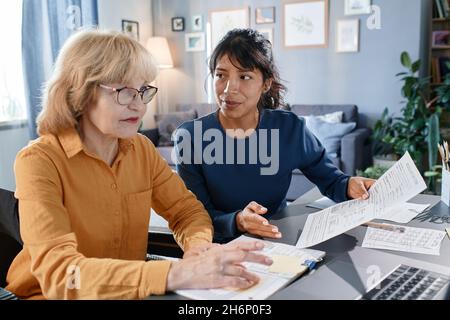 Senior businesswoman in eyeglasses working on laptop with documents at the table together with assistant, they working at home Stock Photo