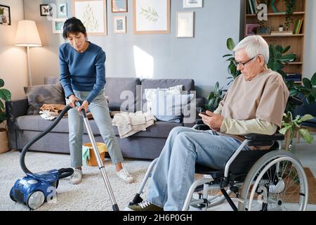 Senior man using mobile phone while housekeeper helping him to clean the room with vacuum cleaner Stock Photo