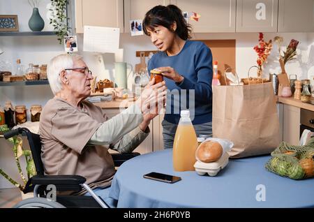 Volunteer delivering food for senior man who using wheelchair, they sitting in the kitchen and discussing food Stock Photo