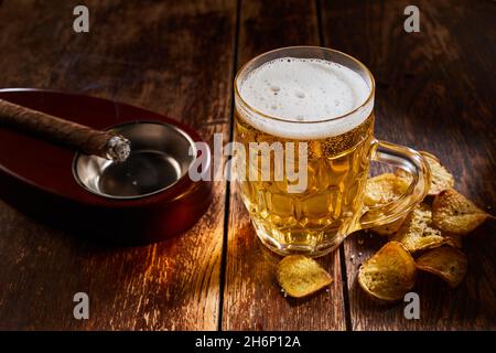 From above of glass of cold lager beer served on wooden table with crispy potato chips and cigar on ashtray Stock Photo