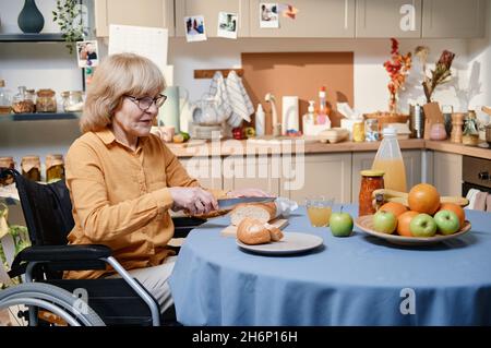 Senior woman using wheelchair sitting at the table and cutting bread, she cooking breakfast for herself in the kitchen Stock Photo