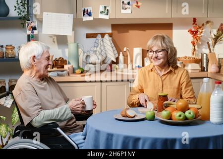 Senior man using wheelchair talking to his wife, she smiling and cutting bread at the table in the kitchen Stock Photo